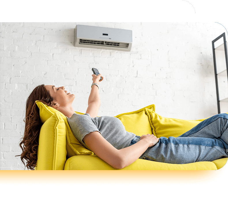 Femme allongée sur un canapé jaune allumant une climatisation murale grâce à une télécommande